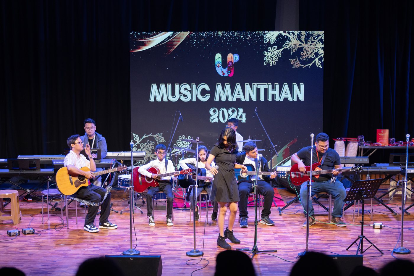 Guitarists seated together on stage with music stands and a drum kit behind them.