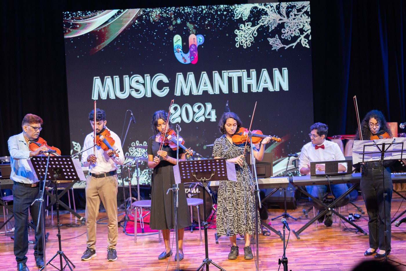 Violinists and instrumentalists performing together with keyboard and guitar on stage.