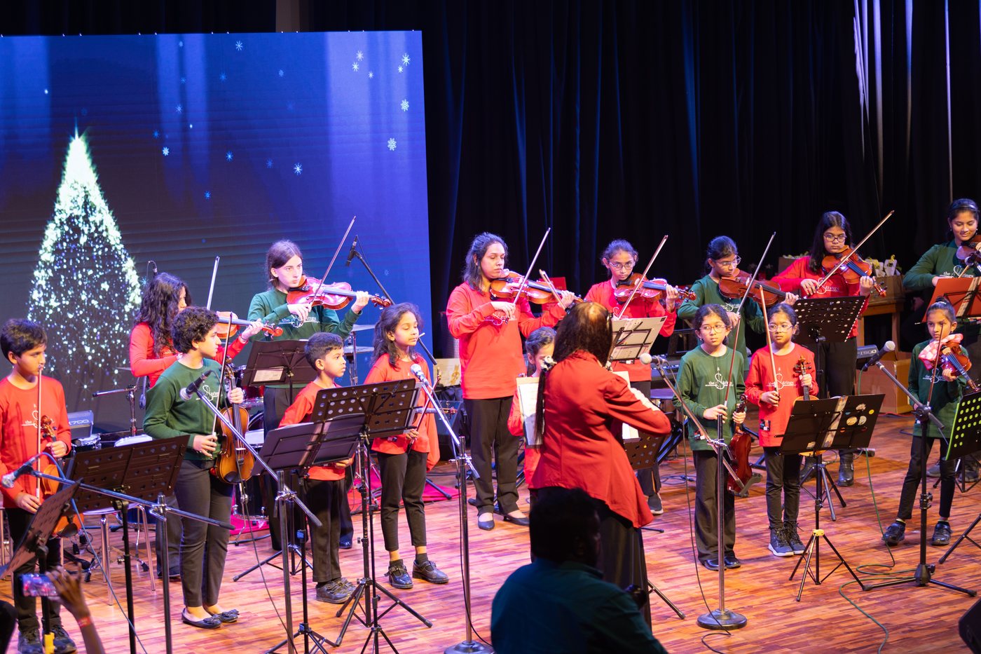 Violin students with bows raised, following a conductor on stage.